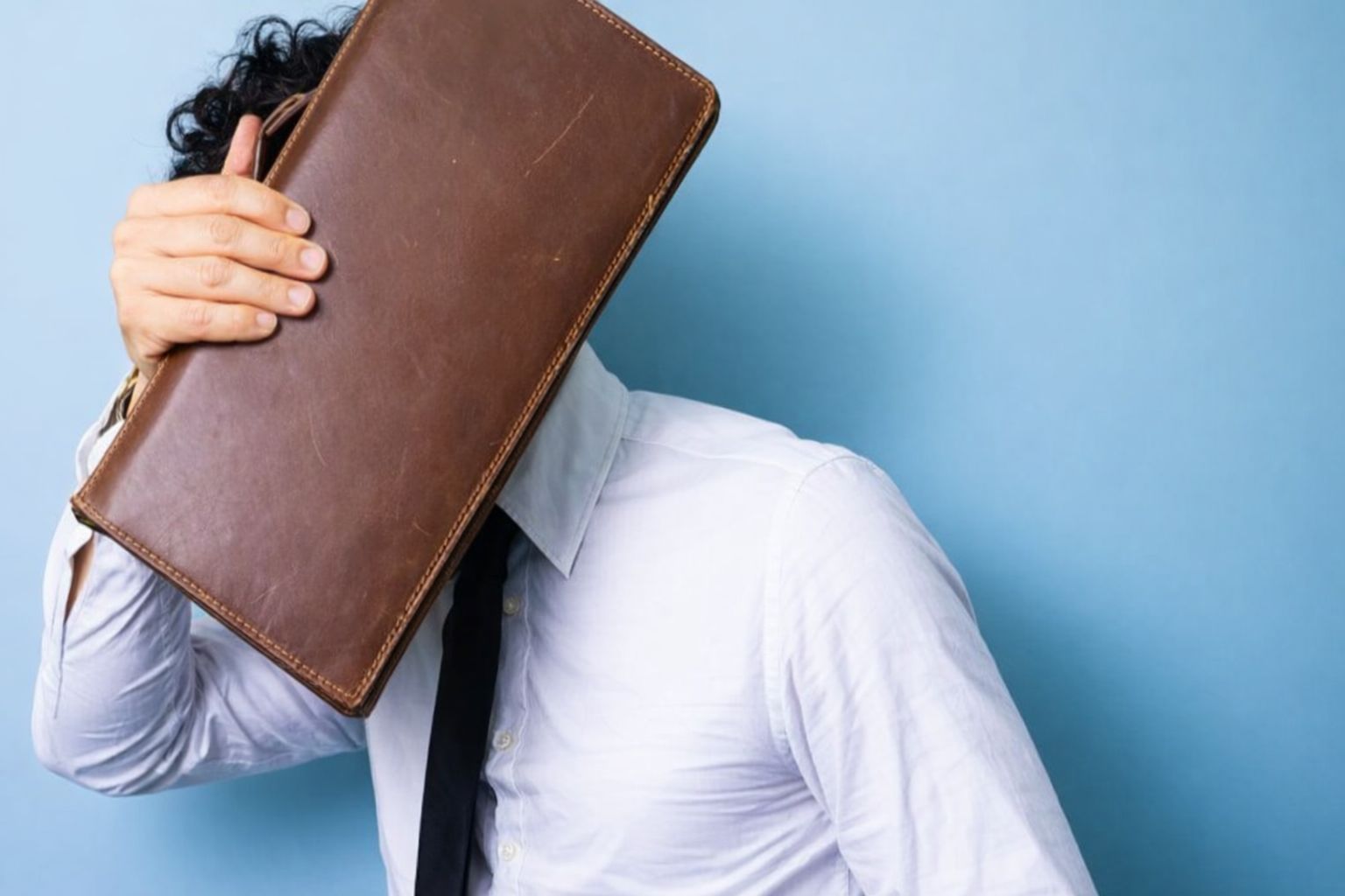 Affärsman gömmer ansiktet bakom en brun portfölj mot blå bakgrund.
